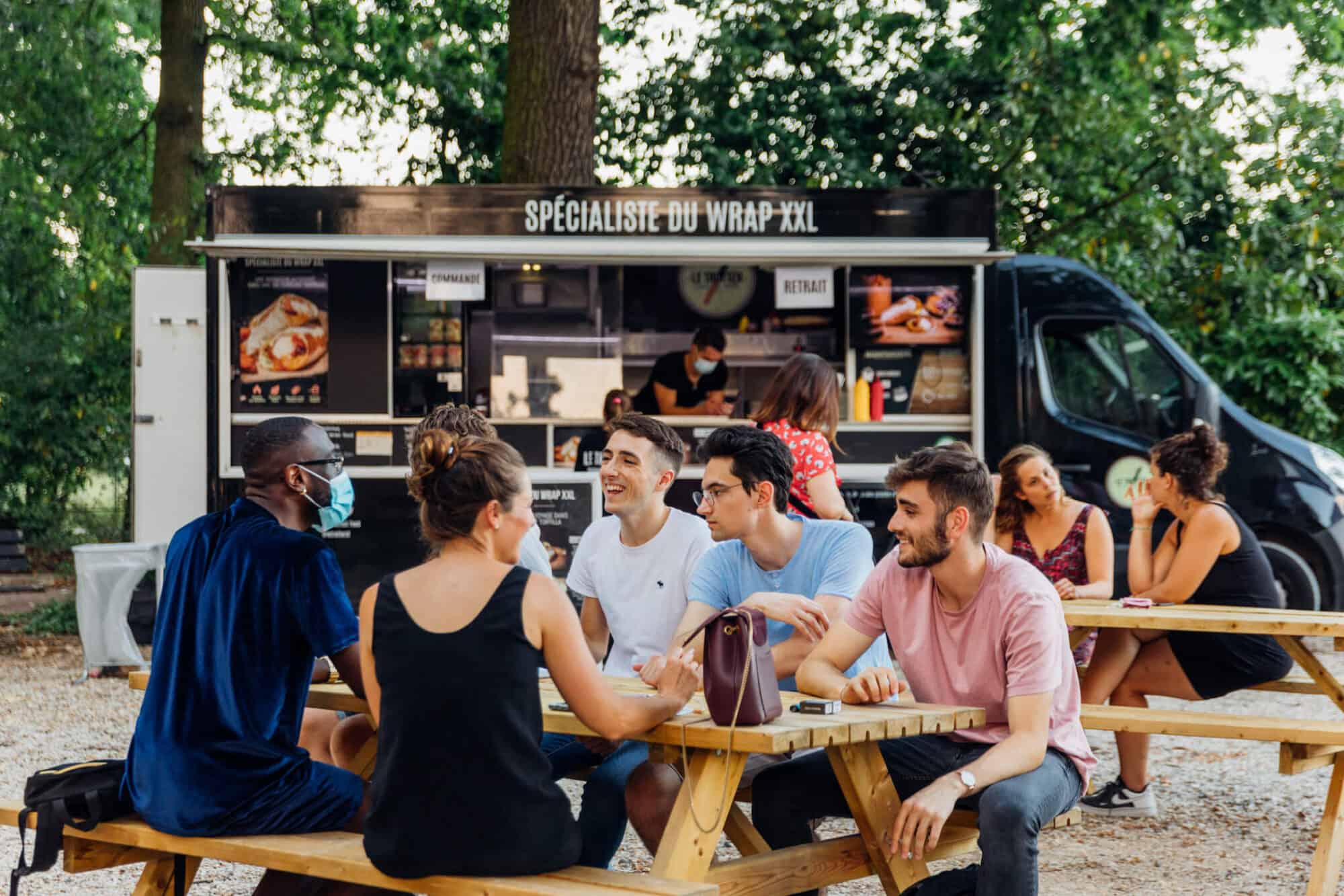 Two groups of people seated at two wooden bench tables in front of a black food truck which has the words 'specialiste du wrap xxl' on it. There is a server in the truck wearing a mask, on his phone. Behind the truck are green trees. 